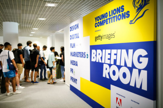 People queuing up next to a yellow and blue board that says Young Lions Competitions